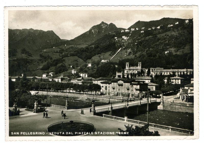 The image depicts a vintage postcard showing the town of San Pellegrino Terme in Italy. The scene is captured from an elevated vantage point, overlooking buildings and streets that are nestled against rolling hills with lush greenery. In the foreground, there's a wide public square featuring pedestrians walking on paths alongside a fountain, bridge crossing over water features, and benches placed around for relaxation. A prominent church or monastery structure stands at the far end of this town square, adding to its picturesque quality.

The text on the image reads "SAN PELLEGRINO - VEDUTA DAL PIAZZALE STAZIONE TERME," indicating that it's a view from Terme Station Square in San Pellegrino. The black and white nature of the photograph suggests it was taken during an earlier time period, likely mid-20th century or before.

Additional information about this image can be found at the referenced URL: https://images.loener.nl/FerroCandilera/full/6730/6730b87489b10e7a26b844a2.jpg. This e