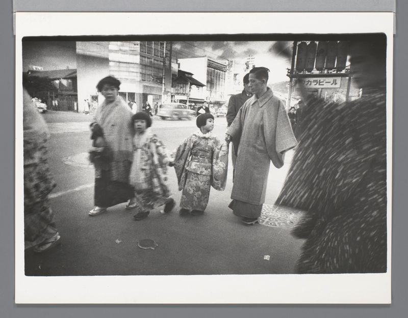 The image is a black and white photograph depicting a family of four on what appears to be an urban street. The setting suggests it may have been taken in Japan, as indicated by the presence of Japanese signage in the background.

Two adults are walking with two children; all members wear traditional attire. The adult leading from left to right seems older than 30 years old and wears a kimono-like garment over casual clothing, holding hands with one child. This suggests they could be parents or relatives responsible for taking care of the children during this outing.

The first child appears younger, possibly around seven years old, dressed in traditional Japanese clothing known as a yukata, which is similar to a kimono but lighter and typically worn during summer festivals such as Bon Odori.

Following closely behind are two other adults. One adult on the right side of the image seems older than 30 or perhaps middle-aged, wearing what looks like a suit jacket over casual clothin