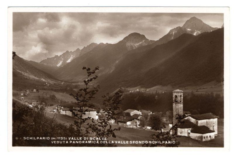 The image depicts a monochromatic photograph of Schilpario, located at an altitude of approximately 1135 meters in the Valle di Scalve region. It captures a panoramic view from above showcasing a picturesque village nestled among rolling hills and towering mountains with their snow-capped peaks piercing through partly cloudy skies.

In the foreground stands a prominent bell tower attached to what appears to be a church or large building, indicating an element of historical significance within this small community. The architecture is traditional European in style, characterized by rustic rooftops and stone facades typical for such settings. 

Surrounding these central structures are smaller buildings with pitched roofs spread across the landscape, creating clusters that signify individual homes and possibly businesses. These settlements suggest a close-knit rural society living harmoniously within this stunning natural environment.

The vastness of the mountains in the background