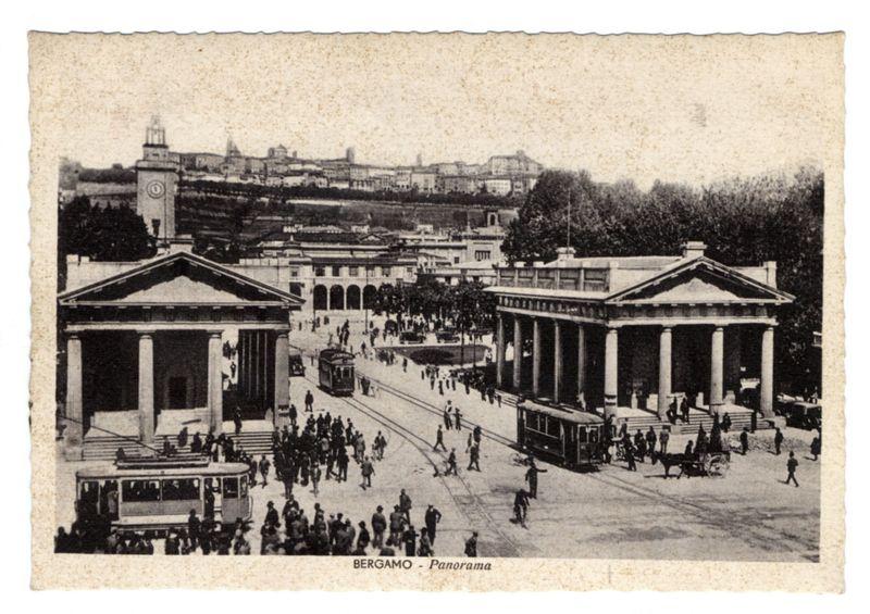 The image depicts a historical black and white photograph of Bergamo, an Italian city. It captures the bustling atmosphere in what appears to be a central square or plaza with classical architecture on either side, including neoclassical buildings with columns. The street is busy with pedestrians walking around and tram vehicles operating on tracks laid out through it.

In the foreground, there's visible wear on the paper-like texture of the photo suggesting age. In the background, various city structures are shown in a panoramic view extending into rolling hills or mountains under a slightly overcast sky. There's a watermark that reads "BERGAMO - Panorama," indicating the location and possibly its historical significance.

The setting reflects an earlier era, potentially late 19th to early 20th century based on the architectural style of buildings, transportation vehicles like trams, and clothing styles visible among people. This image provides a glimpse into Bergamo's urban lands