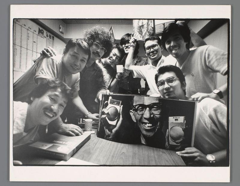 This black and white photograph captures a group of seven individuals, presumably friends or colleagues, in what appears to be an office setting. They are gathered around a table with various items including books, cups, and electronic equipment like cassette players. The central focus is on a large album or book open on the table displaying prominent photographs; one features a man holding up a vintage camera labeled "Nikon," while another depicts a smiling individual wearing glasses. Each person in the photo has a unique expression ranging from smiles to playful gestures such as a hand over their mouth and raised fingers, suggesting a jovial atmosphere. The background includes office elements like a calendar with Japanese characters on it, further indicating that this could be an East Asian workplace or environment. No discernible text provides context for the individuals' identities or specific activities within this image.