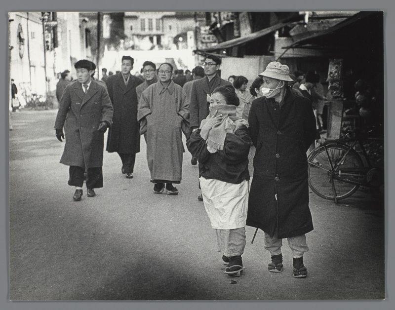 The image depicts a street scene from what appears to be mid-20th century Japan, given the style of clothing and overall atmosphere. A group of people is walking down an urban street; their attire suggests it might be cool weather as most are wearing coats or overcoats with hats for headwear.

In the foreground, there's a man dressed in dark clothes holding what seems to be a child wrapped in cloth on his back, indicating that he could be traveling with family. The second individual walking beside him is older and wears glasses; they have a mask around their chin instead of covering their nose and mouth completely, possibly due to the temperature or local custom at the time.

The background includes other individuals wearing coats who are also in motion on the street. Some buildings can be seen along with what looks like market stalls or shops lining one side of the road. A bicycle is being carried by a person further back from the camera, adding to the sense of movement and daily 