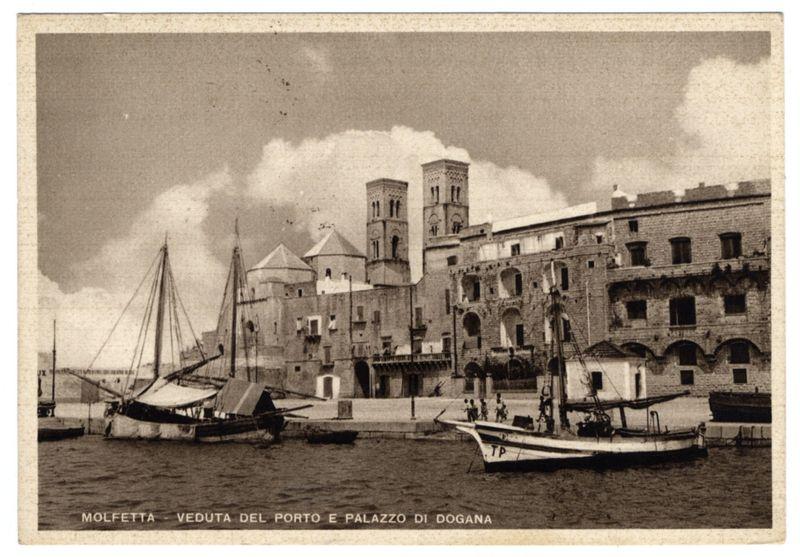 This sepia-toned photograph captures a historical maritime scene, depicting the port of Molfetta with notable architectural structures in view. In the foreground, several boats are moored to the dock and floating near it; some have masts raised while others rest on their hulls. The architecture features Mediterranean styles with arched windows and robust stone construction indicative of coastal defense or commercial buildings from a bygone era.

In the background stands a prominent multi-tiered building with Gothic architectural elements such as pointed arches, ornate detailing along its facade, and tall towers suggestive of historical significance, possibly religious or civic in nature. The sky is overcast, providing diffuse lighting that enhances details without strong shadows. There are no discernible human figures that indicate the time period precisely; however, the style suggests a setting from perhaps late 19th to early 20th century.

The text on the image reads "MOLFETTA - 