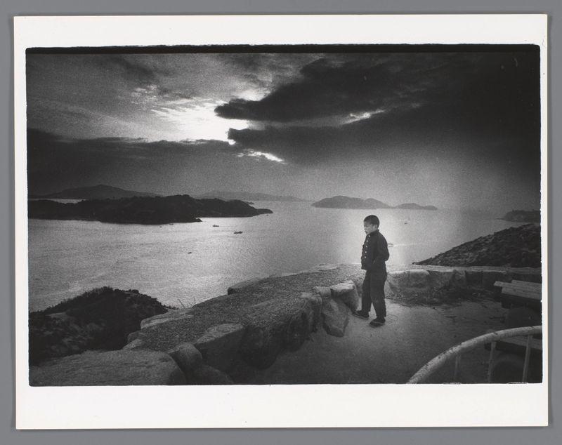 The image is a black and white photograph depicting an individual standing on what appears to be a rugged shoreline. The person, possibly male based on their silhouette and attire, is facing the vast expanse of water with distant islands or landforms in the background under a cloudy sky. The lighting suggests it could be either dawn or dusk due to the soft illumination from behind, creating an atmospheric effect. There are scattered rocks around where the individual stands, adding texture to the foreground. In the distance across the body of water, several small boats can be seen moving gently on the surface. This scene conveys a sense of solitude and contemplation amidst nature's vastness.