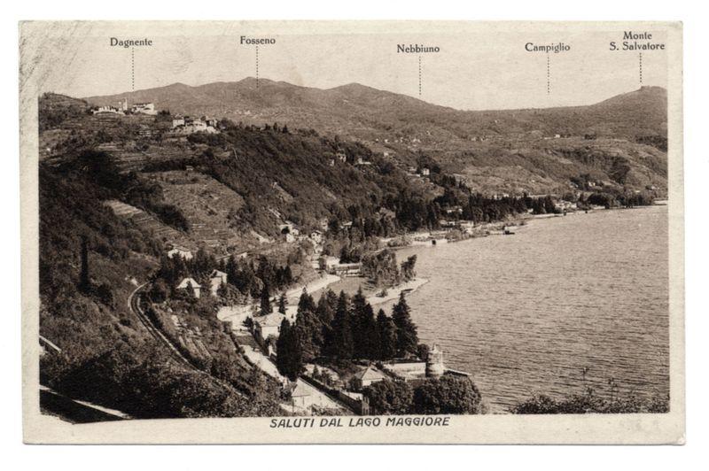 The image is a black and white vintage postcard depicting an aerial view of the Lake Maggiore region. The landscape shows varying elevations with hills dotted by structures, likely buildings or homes, spread out across them. A dense forest covers much of the lower slopes leading down to what appears to be cultivated fields. In contrast, the uppermost areas are more open and feature a few larger establishments that might suggest religious significance due to their elevated position.

A winding road snakes its way through the landscape towards Lake Maggiore which is depicted in calm waters on the right side of the image extending into the distance where it meets the horizon. The lake appears tranquil, indicating either no strong winds or very still weather conditions at the time when this photograph was taken.

Various labels are placed along the top edge and left-hand side identifying different areas such as "Dagnente," "Fosso," "Nebbiuno," among others like "Monte S. Salvatore" on a hill to the far right, indicating geographical markers or notable places in the vicinity of Lake Maggiore.

The bottom of the postcard features prominent text that reads "SALUTI DAL LAGO MAGGIORE", which translates from Italian as “Greetings From Lake Maggiore.” This suggests that the photo was intended for sending warm regards to someone, possibly a loved one or friend who resides near this s [...]