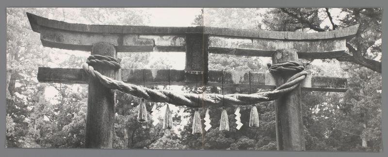 The image displays a black and white photograph of a traditional Japanese torii gate, which is an arch that traditionally marks the entrance to Shinto shrines. The stone structure has two horizontal beams supported by upright posts with curved ends. Draped across these beams are what appear to be paper streamers or flags known as "shide," commonly used in Shinto rituals and festivals for their auspiciousness.

The torii is set against a backdrop of dense forested vegetation, indicating that it may be located within a natural setting such as a mountainous region. The photograph's monochrome palette emphasizes the textures and forms present without the distraction of color. There are no discernible human figures or modern elements in sight; this evokes a sense of historical authenticity.

The image captures the essence of traditional Japanese culture, particularly its religious aspects through Shintoism, with an emphasis on reverence for nature as part of spiritual practice. The photograph seems to be taken from afar due to the lack of perspective lines leading into depth or any indication that it was shot at close range by a lens such as a telephoto.

The specific historical context provided is limited but can suggest several possibilities: Torii gates are typically found in Japan and often commemorate significant events, donations, or dedication. Given this gate's conditi [...]