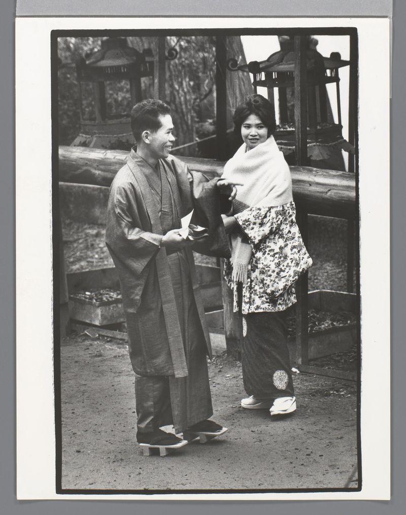 The image is a black and white photograph depicting two individuals in traditional Japanese attire. The person on the left appears to be wearing a kimono, with layered clothing that suggests it might be for colder weather or ceremonial occasion, given its thickness compared to common everyday wear. They are holding what seems like folded paper money or slips of paper towards another individual.

The second person is draped over their shoulders and arms in layers: an outer garment resembling a furisode—a type of long-sleeved kimono often worn during festivals—and underneath appears to be wearing the same layered clothing as seen on the first, possibly indicating a woman's attire. The visible part shows intricate floral patterns.

Both individuals are adorned with traditional footwear known as geta (wooden sandals), which is commonly used in Japan for both formal and casual occasions within Japanese culture.

The background suggests they may be outside at night or under shaded areas due to the darkness enveloping it, with indistinct structures that resemble a park setting. The photo's overall composition reflects cultural elements of Japanese life during what could possibly be an event like New Year celebrations in Kyoto as indicated by additional descriptive information provided about this image.

The photograph has an aged look, which indicates it might have been taken se [...]