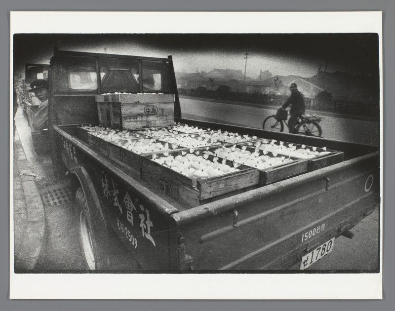 The image depicts a black and white photograph of an old, vintage-style pickup truck filled with numerous cartons that appear to contain food products. The vehicle is parked on the side of what seems like a road or street during twilight hours as indicated by the dimly lit sky in the background.

Inside the bed of the truck are wooden crates labeled "金福" which translates to "Golden Fortune," and there's also text indicating prices, such as "1500円", meaning 1500 yen. The vehicle has a visible license plate with Japanese characters on it. There is at least one person standing next to or inside the truck, partially obscured by its structure.

In the background, a cyclist can be seen riding away from the viewpoint of the photo. Buildings and some faint street infrastructure are discernible in the far distance under an overcast sky. The overall setting suggests this might have been taken during a time when Japanese cities were developing or modernizing their streetscapes.

This image is associated with Ed van der Elsken, as mentioned in the additional information provided which references his work "Vrachtwagen in Japan" (Truck in Japan) from 1959-60.
