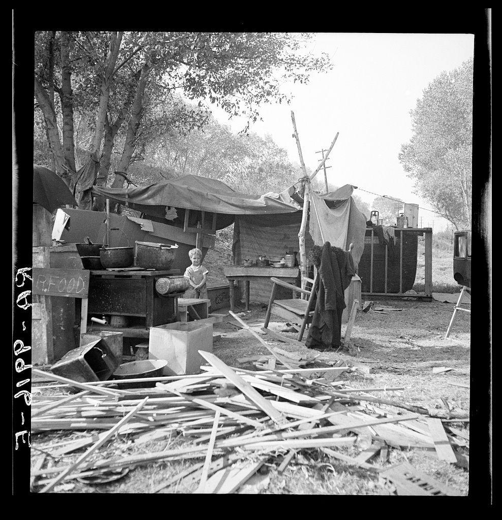 The image depicts a black and white photograph of what appears to be an impoverished family living in temporary camp conditions. A child is standing near the center, possibly engaging with something or someone outside of view. The environment around them looks unkempt with scattered wooden planks on the ground, suggesting disrepair or makeshift shelter construction.

In the background, there are various items and structures including a tent-like covering supported by poles, an old-fashioned stove with pots placed on top, and miscellaneous household items such as kerosene lamps and containers. The overall scene conveys hardship and lack of resources among this family during what appears to be their living situation in a campsite.

This photograph is identified as being from the American River Camp near Sacramento, California, illustrating the plight of destitute families possibly related to rural or economic challenges faced by Americans at that time.