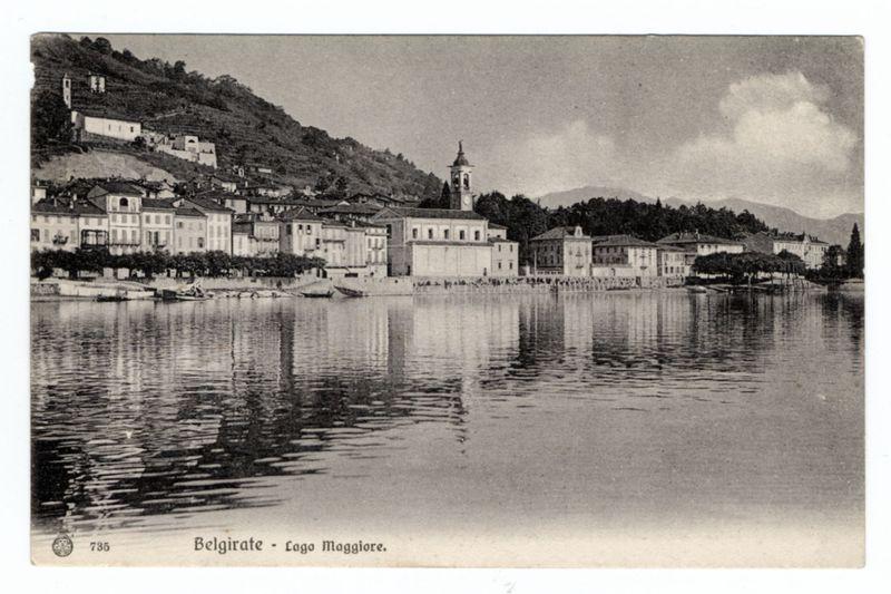 The image is a black and white photograph depicting the lakeside village of Belgirate, located on Lake Maggiore in Italy. In the foreground, calm waters reflect the structures along the shoreline. The middle ground shows an array of buildings with distinct European architecture, possibly residential or public edifices. Prominently positioned among these structures is a church with its bell tower standing tall against the partly cloudy sky.

The background reveals rolling hillsides dotted with vegetation and what appears to be vineyards, indicative of wine production in this region. The overall atmosphere suggests a serene and picturesque locale popular for tourism or as a residential area nestled within nature's embrace. There are no visible modern elements that might date the photo beyond its historical context; it could potentially range from late 19th century to early mid-20th century, judging by architectural styles and lack of contemporary infrastructure.

The image is labeled