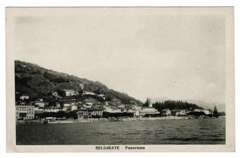 The image displays a historical black and white photograph of the town of Belgirate, located on the shore of Lago Maggiore in Italy. The scene captures multiple buildings with varying architectural styles nestled against rolling hills that descend into the lake's edge. Prominent among these is what appears to be St. George Church at its highest point, standing out due to its white façade and bell tower, surrounded by dense greenery. In front of this townscape lies a body of water indicative of Lago Maggiore with subtle ripples on the surface. The image bears text that reads "BELGIRATE - Panorama" in Italian at the bottom center, suggesting it may be an old postcard or albumen silver print from the late 19th to early 20th century based on its style and quality of printing. There's a faded watermark across the top right corner with what looks like "P. Cadeddu", potentially indicating the photographer's mark.