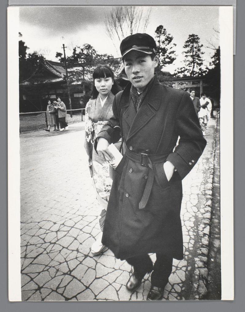 The image is a black and white photograph depicting two individuals standing on what appears to be a paved path. The person in the foreground, likely male based on appearance, wears a dark coat with a belt, trousers, shoes, and has a cap on his head. He holds something small in one hand and looks directly at the camera with a neutral expression.

The individual behind him is wearing traditional Japanese attire known as a kimono adorned with floral patterns, suggesting they are dressed for cultural or ceremonial purposes. The setting appears to be an outdoor area near traditional Japanese architecture; there's evidence of trees and structures that resemble buildings typical in historical areas such as temples or shrines.

In the background, several other people can be seen walking along what looks like a sidewalk made from irregularly shaped stones, indicating it might be part of a temple complex. The overall atmosphere suggests this could have been taken during an event where tradi