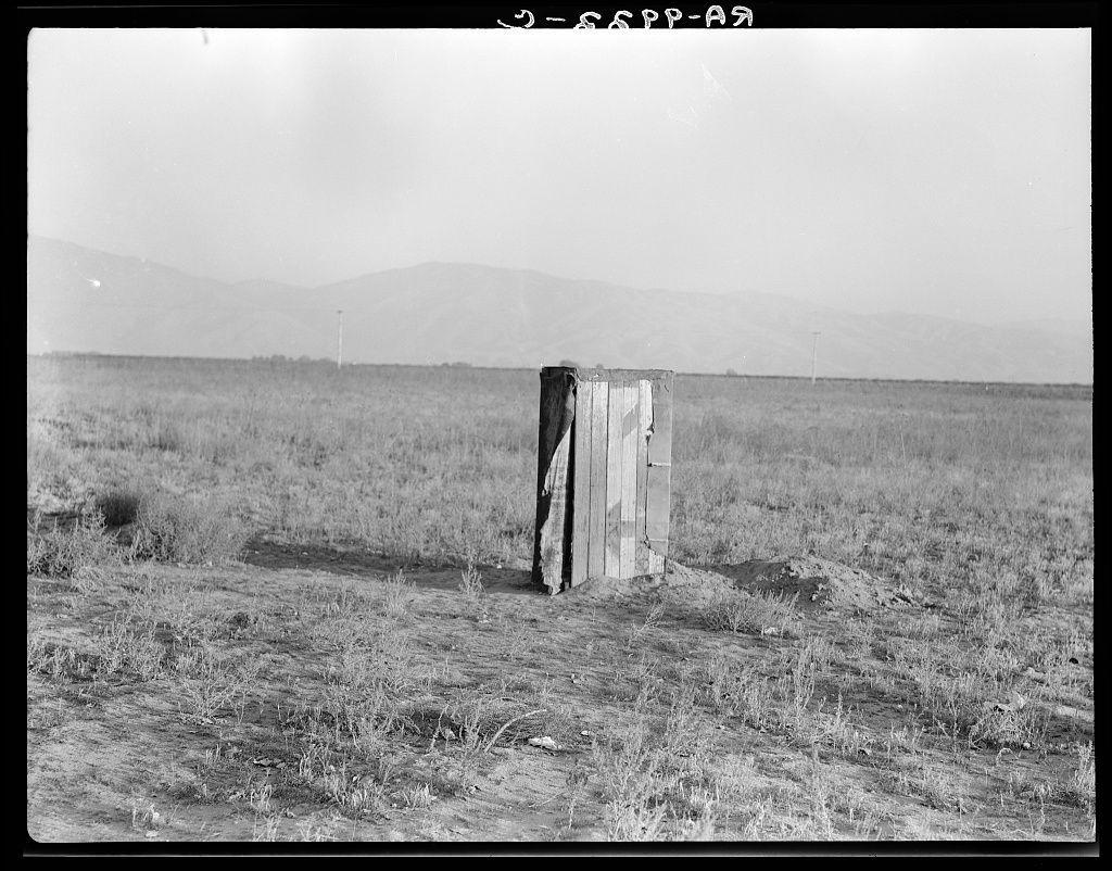 The image shows a solitary wooden structure in an expansive field. This appears to be an improvised toilet or sanitation facility, indicative of conditions faced by migratory workers during agricultural seasons as mentioned in the provided source information. The photograph is black and white with high contrast, accentuating shadows and texture contrasts between sunlight-exposed areas and shadowed regions. In the background, there's a vast open field leading up to distant mountains under an overcast sky. No human subjects are visible within this shot, focusing entirely on the sanitation setup amidst rural or semi-rural terrain.