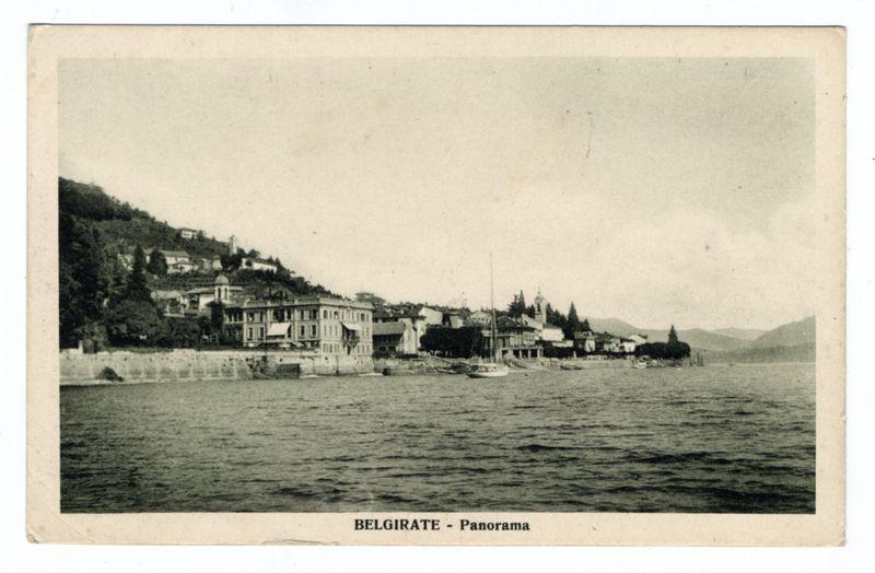 The image displays a vintage black and white postcard titled "BELGIRATE - Panorama," depicting the scenic waterfront of Belgrade, situated by a body of water. The photograph captures an array of European-style buildings lining the shore on one side and rolling hills with dense vegetation in the background. Among these structures, there are several larger homes or possibly hotels prominently featured alongside smaller residential houses. A fortified wall borders part of the shoreline, suggesting historical significance to the area.

Several boats float gently on the water's surface, including a small boat near what appears to be a pier and another closer to one of the prominent buildings by the shore. The calmness of the waters hints at peaceful weather conditions during the time when this photo was taken.

The sky above is overcast with soft clouds diffusing sunlight, giving an overall serene ambiance to the scene. No people are visible in this image, which focuses on capturing the