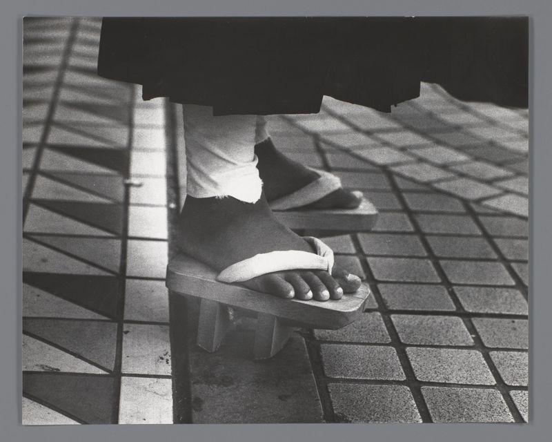 The image captures a close-up of someone's feet wearing traditional Japanese geta sandals. The person appears to be standing on tiled flooring, and the shadows cast by their legs create geometric patterns across the surface. The monochromatic tone of the photograph emphasizes contrast between light and shadow, giving it a dramatic effect. This style is often associated with documentary or street photography that focuses on capturing everyday life in Japan during the 1980s.