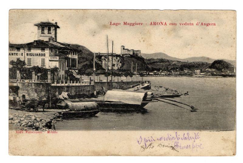 The image depicts a vintage postcard from Lago Maggiore, specifically Arona with views of Angera. The photograph is in black and white, giving it an aged appearance consistent with historical photographs. In the foreground, there are several boats moored on calm waters near the shore; some have covers over them while others show their wooden hulls exposed. A small rowboat rests nearby without a cover.

In the middle ground, we see multiple masts rising from what appears to be docks or piers extending into the lake. The background features an elaborate building with classical architectural elements on its facade labeled "Ante e Bigliardo." Behind it is another structure perched atop a hillside overlooking the waters and landscape beyond.

The distant hills create a backdrop of natural beauty, with terraced fields indicative of agricultural use. Several people are visible in small groups near some structures along the lakeside, giving life to this serene setting.

Atop the postcard