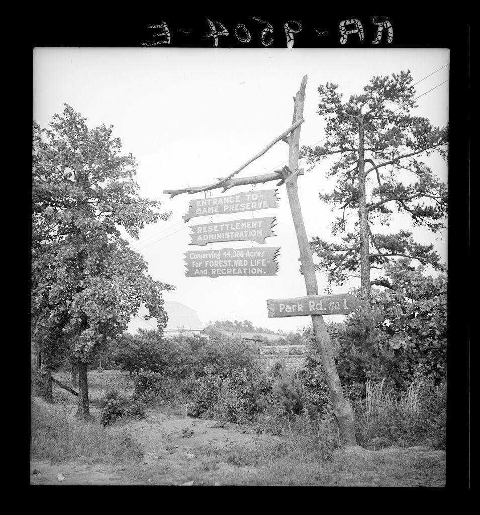 The photograph is a black and white image showing multiple wooden signposts in an outdoor setting, likely part of a rural landscape. The signs provide directions to various locations including the "Entrance To Game Preserver," "Resettlement Administration," among others, indicating their historical significance related to land use for resettlement projects or game preservation during times such as after World War II when many were displaced.

The signpost with directional arrows is in focus and seems like it's meant to guide visitors. It indicates directions towards various administrative offices possibly located nearby the area that was part of a larger project aimed at providing homes, jobs, land grants for farmers who had been displaced by war or economic downturns.

In the background, there are trees and shrubs indicating an outdoor environment with natural vegetation which suggests this photo could have been taken in Georgia as per the provided link. The landscape is not urban