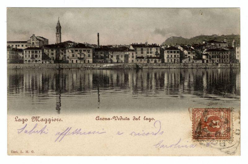 The image is a sepia-toned postcard featuring an old photograph of the town Arona in Lago Maggiore, Italy. The scene captures the tranquil waters reflecting buildings on their banks with one prominent church tower and several smaller structures. There's handwritten text at the bottom left corner that appears to be signatures or annotations, including "Esel. L. M. G." possibly indicating the sender, followed by additional names in cursive script which could refer to recipients. At the top right is a red postage stamp with an image of what seems like a queen and bears Italian text within it; however, specific details are not clearly readable due to size constraints on this description. The overall condition suggests age and usage typical for vintage postcards from historical periods.