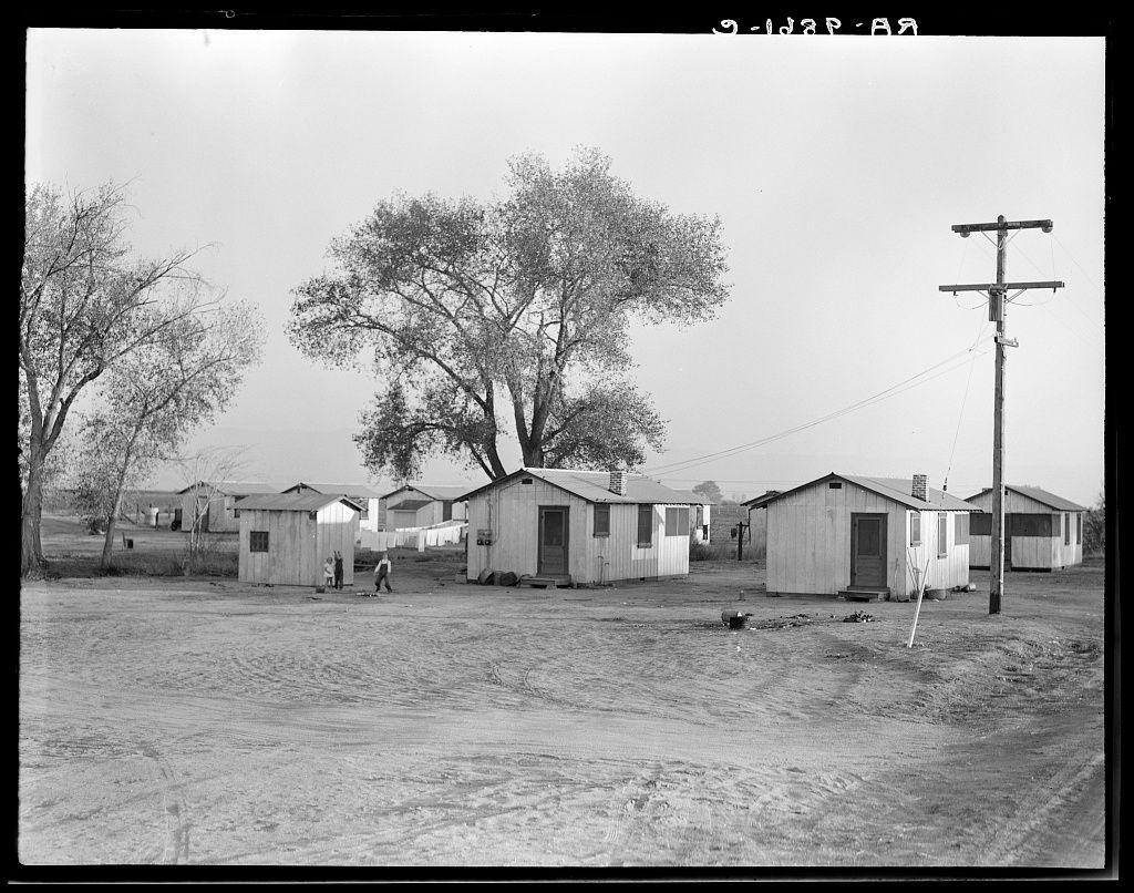 The image is a black and white photograph depicting an outdoor scene with several small wooden cabins arranged in what appears to be a makeshift settlement. The ground seems unpaved, possibly dirt or gravel, indicating a rural setting. There are multiple telephone poles scattered around the area, suggesting some level of infrastructure development but not elaborate by any means.

In front of one cabin, there's an adult and two children; it is unclear whether they live in this particular structure or if they're just passing through. The trees have sparse foliage, possibly indicating a season where leaves are scarce such as late fall or winter, which adds to the overall austere atmosphere of the scene.

The condition of these structures implies that they may not be intended for long-term habitation but rather temporary housing solutions. This setting could potentially represent an instance from past socioeconomic challenges like the Great Depression when many people faced displacemen