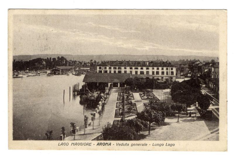 The image depicts a historical postcard showing an early 20th-century view of the lakeside promenade in Arona, situated on Lake Maggiore. The scene captures several key elements: 

- A large building with a flat roof and multiple windows dominates the center-right portion of the frame.
- In front of this structure is a paved area lined by rows of trees and evenly spaced lampposts, leading towards the waterfront where boats are docked along wooden piers.
- The shoreline features neatly arranged lamp posts and tree-lined paths, suggesting well-maintained public spaces typical for leisurely strolls or social gatherings.

The water body reflects the hazy sky above with no visible waves. In the background, a landscape of hills is faintly discernible under an overcast sky, contributing to the tranquil atmosphere conveyed by this sepia-toned photograph.
This image does not include any identifiable individuals and focuses on depicting the general layout and ambiance of Arona's waterfron