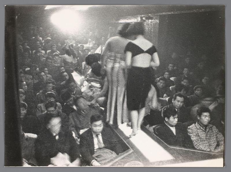 The image is a black and white photograph capturing the vibrant atmosphere of what appears to be a live performance or show. In the foreground, two individuals are walking on stage; one person is wearing an outfit with a large hemline that exposes their legs while another individual in dark clothing accompanies them. The audience surrounds this scene closely, seated tightly together with varied expressions ranging from attention and engagement to disengagement.

The lighting focuses strongly on the performers, creating high contrast between light and shadow within the crowd. Some audience members are captured mid-motion or engaged in conversation. The photograph has a candid quality, suggesting it was taken spontaneously rather than posed for. This evokes an authentic sense of the event's liveliness and immediacy.

Further information about this image can be found at Nachtclub of theater in Japan by Ed van der Elsken (1959 - 1966), with a reference to a specific photograph number, 