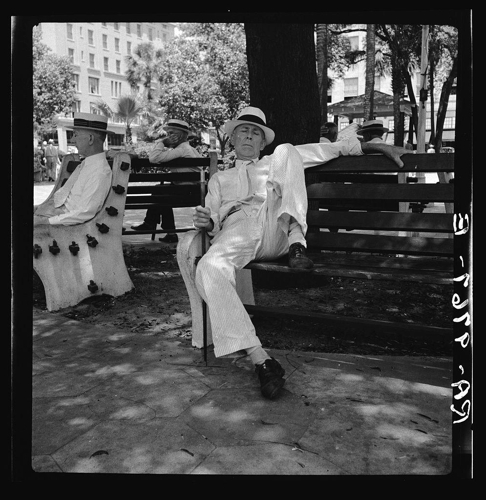 The photograph depicts three individuals seated on benches within an outdoor setting that appears to be a park. The person in the foreground, likely female based on clothing and hairstyle, is leaning back with one leg crossed over her knee while resting against a bench's armrests. She wears striped pants and a white blouse or dress suit paired with dark shoes. Her hat sits slightly tilted as she gazes off to the side.

Behind this individual are two other people seated separately; both appear elderly based on their attire, which includes hats consistent with styles from past decades. The person behind her leans forward while gripping what seems like a cane tightly in hand and is dressed similarly in light-colored stripes or patterns.

The environment suggests an urban park setting during the daytime under clear skies due to shadows cast upon the ground. Trees line the background alongside buildings, possibly indicating that this park exists within a cityscape. The image's black-and