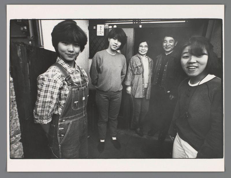 The image is a black and white photograph featuring five individuals, seemingly posing for the camera. From left to right: one person wearing checkered overalls with rolled-up sleeves stands confidently with hands in pockets; beside him are two younger-looking males standing back-to-back, one slightly turned towards the photographer with his arms by his side, both dressed casually in sweaters and jeans. The next individual is a middle-aged male with glasses smiling at the camera, wearing what appears to be workwear including an apron over casual clothes, suggesting he may have been working or on his way to do so; lastly, there's another young female facing directly towards the camera with a broad smile, her hair pulled back. Behind them is a backdrop that seems like an indoor setting, possibly public transport or a small establishment given what looks like part of a bench and some architectural details in the background. There are signs of wear on the image itself, such as creases and 