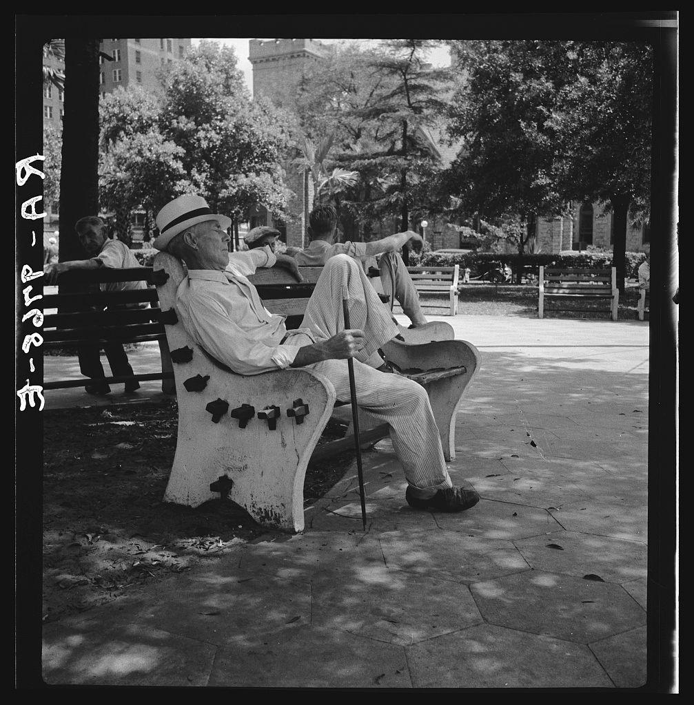The photograph depicts an elderly individual seated on a concrete bench in what appears to be a park setting. The person is dressed casually, wearing a wide-brimmed hat and glasses, with their legs crossed as they rest one foot atop the opposite knee while holding onto a cane for support. Several decorative elements resembling small crosses are affixed along the edge of the bench on which this individual sits.

In the background, other park-goers can be observed sitting or walking by, suggesting that it's a public space where people gather to relax and spend time outdoors. The trees provide shade, indicating either early morning or late afternoon light conditions as there is no direct sunlight falling directly upon the subjects in view. The image conveys a sense of leisurely downtime within an urban environment.

The photograph itself carries signs of age with its black-and-white format, which hints at it being taken during the mid-20th century. Additional context or information ab