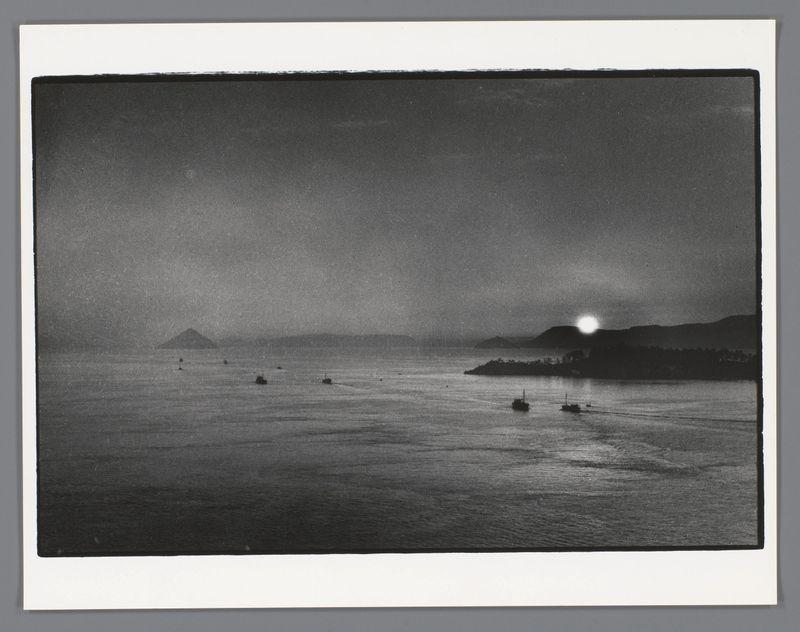 This image depicts a tranquil seascape at dusk or dawn, captured in black and white. The horizon is dominated by the silhouette of an island with what appears to be light activity on its shore, possibly indicating settlement or civilization. In contrast, another larger landmass looms in the background under a sky that transitions from dark near the top to lighter shades closer to the sea's surface.

Floating gently across this vast expanse are several small boats, their presence adding life and scale to the scene without disrupting its serene atmosphere. The water is calm with minimal disturbance except for the gentle ripples created by the moving vessels. There isn't a clear view of any land nearer than the silhouette in the distance; instead, there's an open sea that stretches far into the horizon.

The overall mood conveyed by this photograph suggests quietude and contemplation, as if inviting viewers to reflect on their own experiences or emotions connected with solitude at lar
