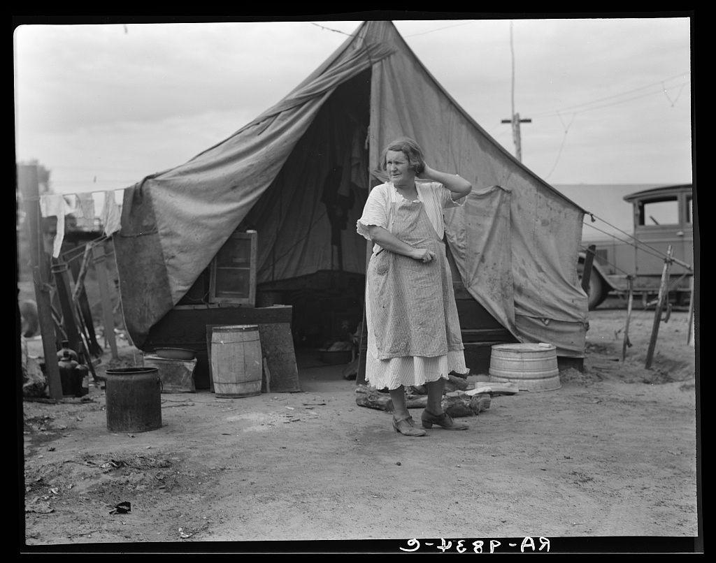 The image depicts a woman standing outside of a makeshift tent structure. The setting appears to be an outdoor, possibly temporary living area with various objects scattered around such as barrels and what seems like agricultural equipment or tools in the background. It is rendered in black-and-white, suggesting it might be from historical records or archival photography.

This photograph likely captures a moment during the Dust Bowl era when migrant workers traveled across states searching for work opportunities, often setting up temporary encampments along their journey. The woman's attire and the overall scene reflect the austere conditions faced by many migrants of that time period in agricultural regions like California.