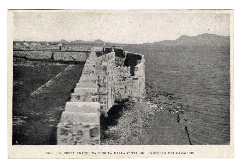 The image is a black and white photograph depicting the historical remains of what appears to be part of an ancient wall or fortification. The structure consists of tall, weathered stone blocks that run alongside a body of water with calm waves visible in the background. This suggests it could have been situated on coastal terrain during its time of use.

In the distance, hills or low mountains can be seen under a pale sky, indicating an open landscape beyond the immediate vicinity of this ruinous site. The photograph has text at the bottom which reads "COO - LA COSTA ANATOLICA VEDUTA DALLA CINTA DEL CASTELLO DEI CAVALIERI," suggesting that this location is in Anatolia and viewed from within Castle Cavalieri's ramparts.

The quality of the image suggests it might be quite old, possibly a postcard or an early photographic print. The condition of the stones indicates significant erosion over time, likely due to weathering effects near the sea.