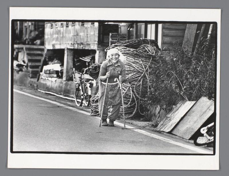 The image depicts an elderly woman standing on a street. She is smiling and wearing crutches in one hand, suggesting she may have mobility issues or be recovering from surgery. The setting appears to be rural with natural vegetation visible behind her and some wooden structures that could be part of farm buildings or storage areas for agricultural supplies.

Behind the woman are large bundles of what looks like rope or nets stacked against a fence, indicating this area might be used for storing fishing gear or similar equipment commonly found in coastal regions. To her left is an old bicycle leaning on one of these bundles, further emphasizing the rustic and utilitarian nature of the scene.

The image conveys a sense of resilience and daily life in what could potentially be a remote community where manual labor plays a significant role. The overall composition captures candidness and authenticity without any apparent staging or artificiality.