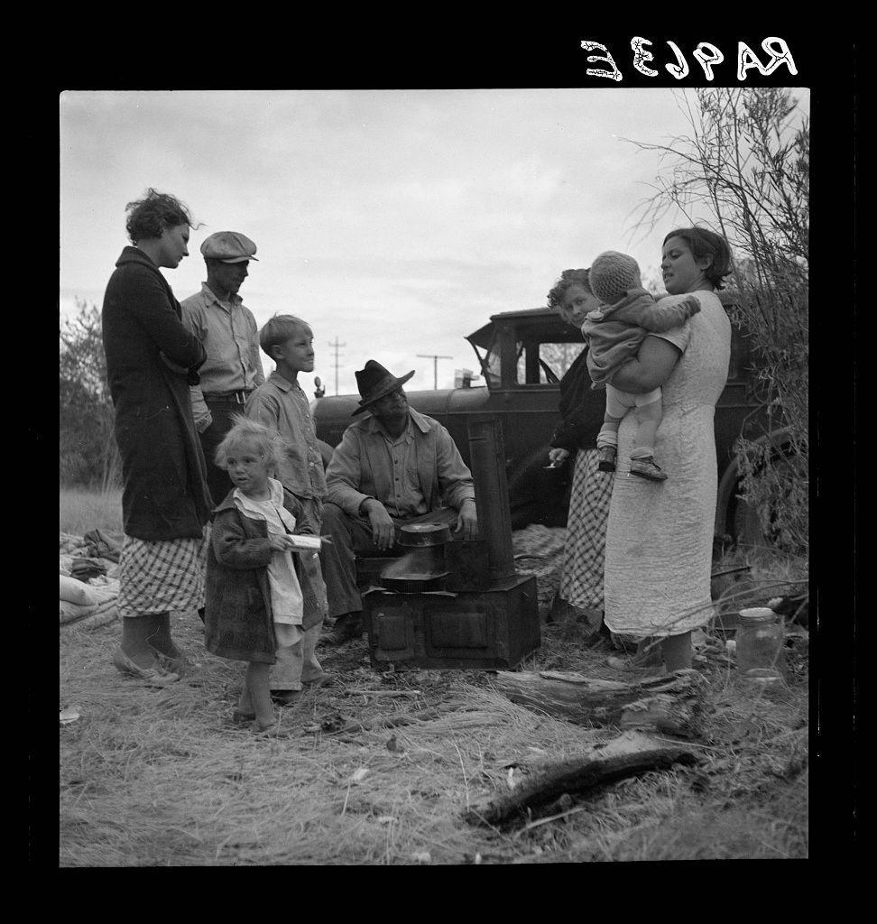 A black and white photograph depicting a group of eight people gathered around an old-fashioned cooking stove outdoors. They are dressed in modest, worn clothing suggesting hardship or poverty; many wear hats and coats indicative of cold weather. The men appear to be older with some wearing caps while the women have their hair uncovered except for one who is holding a child close. Children accompany them; they seem well-cared-for despite the circumstances as indicated by clean clothes on at least two young boys, possibly brothers given similar attire or accessories such as hats and coats that match those of adults around them.

The setting looks rural with sparse vegetation and an overcast sky, suggesting it might be a windy day which is typical in areas affected by droughts like Dust Bowl regions. The photograph seems to capture the essence of people displaced due perhaps to circumstances beyond their control (like severe weather events), as indicated by text written above them like