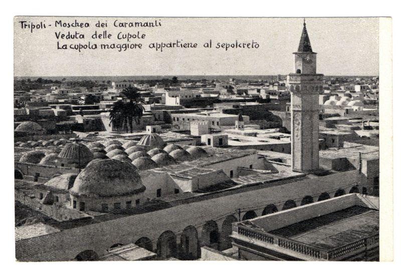 The image shows a historical black and white photograph depicting the city of Tripoli, with prominent Islamic architecture. It features multiple domed structures, likely part of mosques or religious buildings known as 'cupolas', spread across an urban landscape. The largest dome is pointed out to belong to a mausoleum (sepolcreto). In the foreground stands a tall minaret with a clock face visible near its top, suggesting it's associated with one of the mosques in the scene. Text overlaid on the image indicates locations and features such as 'Tripoli - Moschea dei Caramanli' for Tripoli, referring to the Minaret of the Great Mosque (la cupola maggiore appartiene al sepolcreto), which translates from Italian to indicate that one or more domes belong to a mausoleum. The style and condition suggest it is an old photograph possibly dating back several decades ago when such postcards were popular souvenirs for travelers.