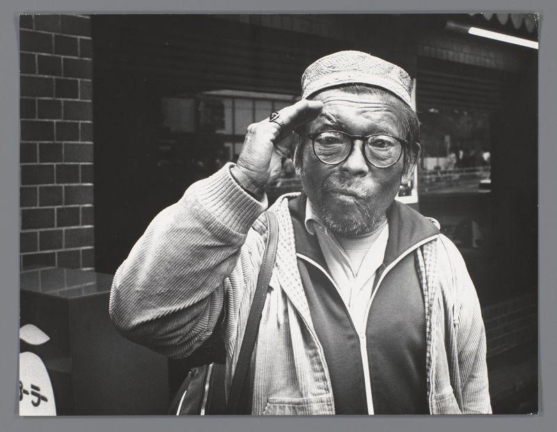 This black and white photograph features an older man saluting with his right hand. He wears glasses, a patterned hat, layered clothing including a vest over a shirt, and carries what appears to be a shoulder bag on the left side of his body. The background is out of focus but shows urban elements such as brick walls and possibly storefronts or signage in Japanese characters, indicating an outdoor setting likely within Japan. His expression is earnest, with visible wrinkles around his eyes suggesting deep thoughtfulness or concentration. There's no additional context provided about this man beyond the visual details captured by the photograph itself.