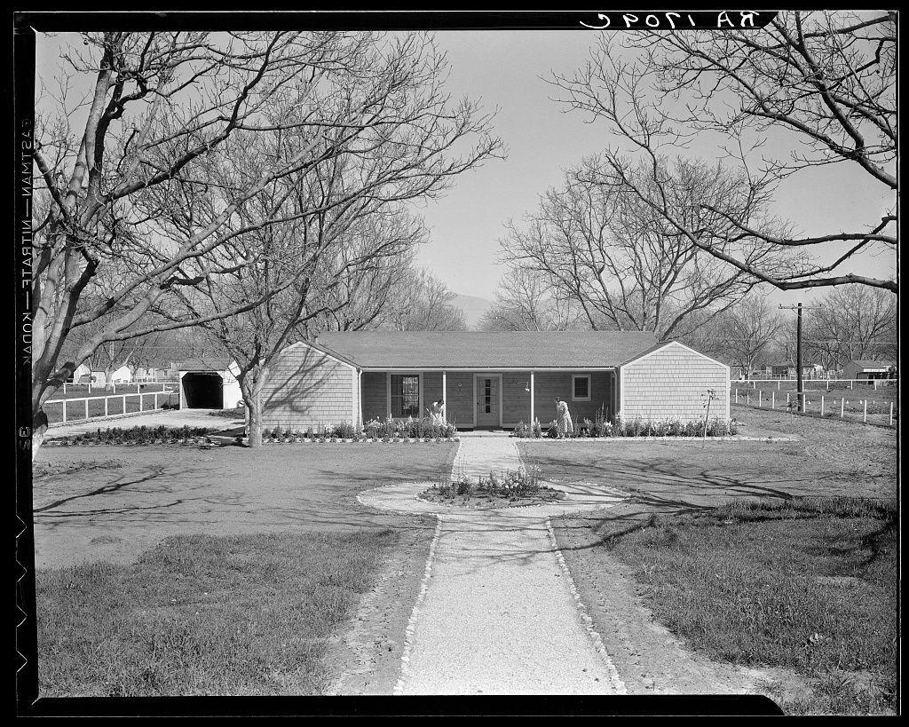 The image depicts a rural landscape featuring a modest, two-bedroom house with an attached garage. The structure is single-story and has wooden siding. It's situated on what appears to be farmland or open pastureland, as indicated by the presence of fences surrounding the property.

A tree-lined path leads up to the front door of the home, where we see two individuals: a woman who seems engaged in gardening activities near flower beds with various plants and flowers, and another person standing at the porch entrance. The setting is devoid of snow or signs of winter weather but suggests an early spring season due to leafless trees.

The photo's black-and-white tone hints that it could be from a historical context, possibly mid-20th century given its style and grayscale presentation. There are no explicit indications of modern elements such as streetlights, cars, or contemporary clothing styles visible in the scene. The environment is tranquil with minimal activity outside, emphasizi