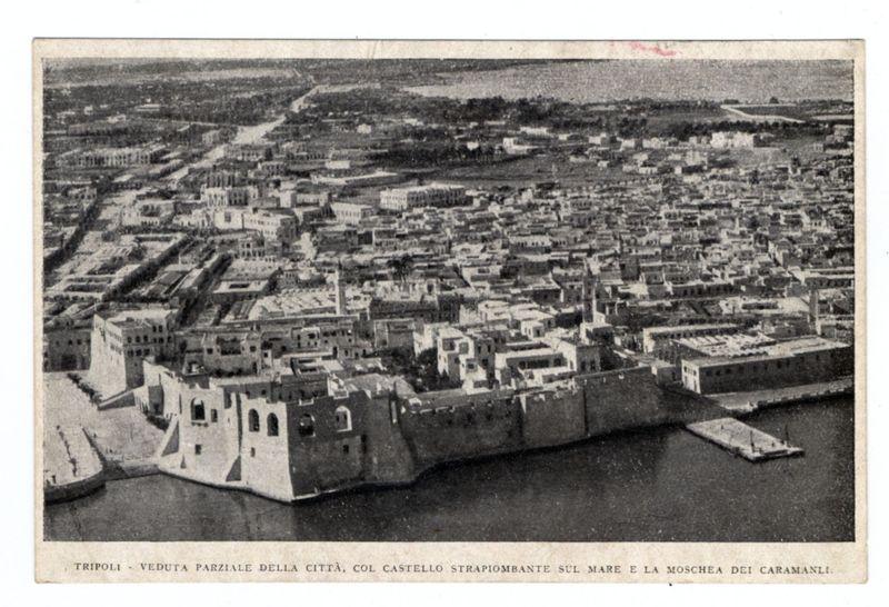 The image depicts a historic aerial view of Tripoli, specifically highlighting the city walls and harbor area known as Veduta Parziale Della Città. The photograph is in black and white, suggesting it's from an earlier time period when color photography was not common or used for such representations. Visible are fortified structures resembling Castello Strapiombante (meaning "the sinking castle"), a nickname likely due to its location at the edge of the water where waves can cause parts of the structure to appear submerged during high tide, as well as Moschea dei Caramanli, which is presumably one of Tripoli's mosques. The urban layout shows tightly packed buildings with uniform architecture typical for cities in this region and era. Notable features include a large harbor area leading up to these walls, hinting at the city’s historical importance as an economic center possibly due to its maritime activity given its coastal position.