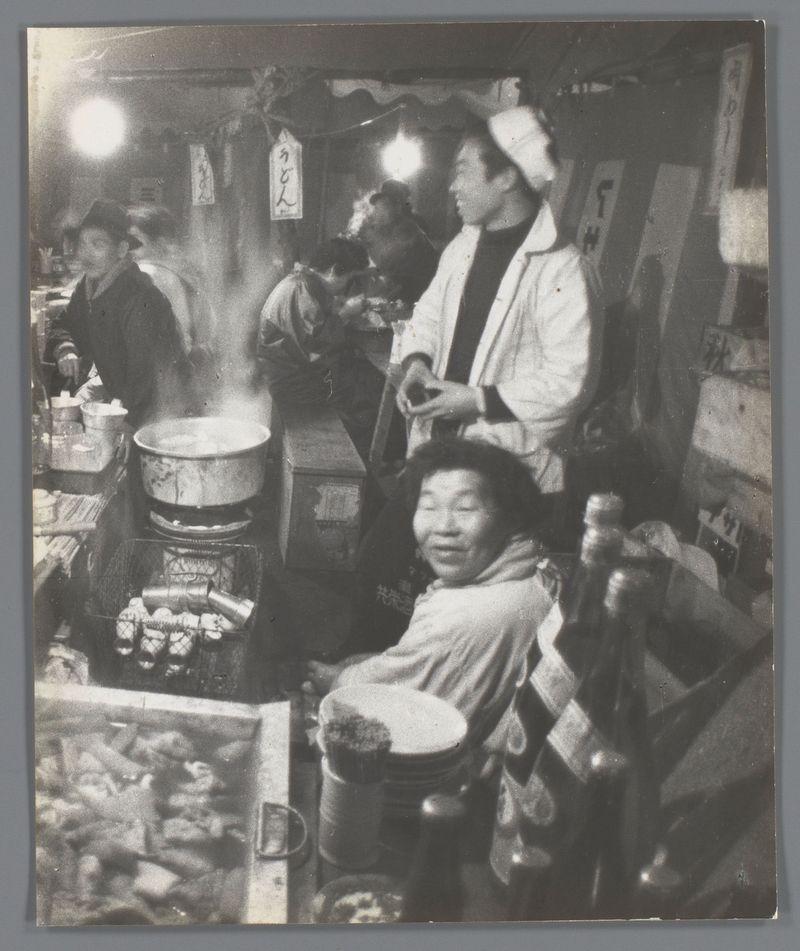 The image depicts an indoor scene, likely a street food stall or small restaurant in Japan. It's captured from behind the counter where various cooking utensils and ingredients are displayed, with steam rising indicating active cooking.

Two individuals wearing white hats suggest they might be chefs or cooks engaged in serving customers or preparing food. In front of them is another individual who appears to be a customer; their focus seems directed towards something outside the frame.

Behind these figures, there's an array of signage and decorations that are typical for such eateries, adding character to the environment. The lighting casts strong contrasts with some areas well-lit while others remain in shadow, enhancing the dynamic feel of the setting.