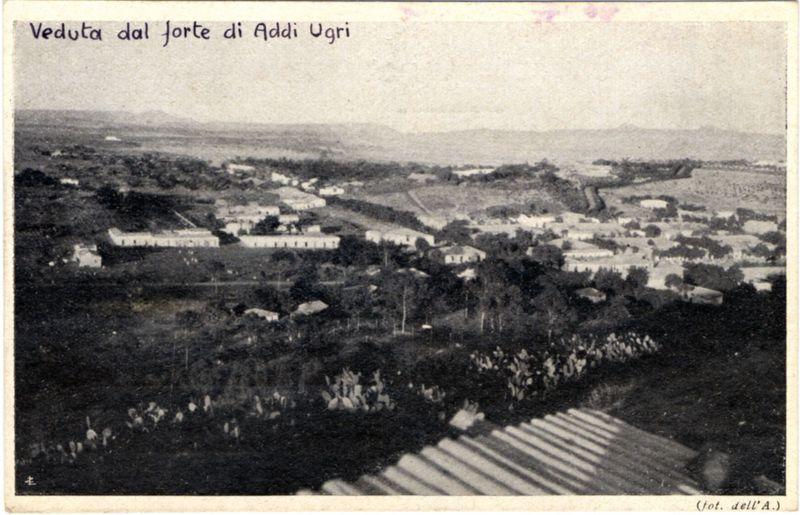 The image is a vintage black and white photograph depicting an aerial view of the Forte di Addi Ugri. It shows various buildings, including what appears to be barracks or administrative structures with flat roofs, situated in a semi-rural landscape. The area around these buildings includes open fields, some sparse vegetation, and scattered trees, suggesting it might have been used for agricultural purposes or as part of an industrial complex during its time.

In the foreground, there is a significant group of people gathered together on what looks like an earthen embankment leading down to a wooden pathway. The crowd seems large enough to suggest some kind of event or assembly was taking place at that moment. In contrast, the upper portion of the image shows more distant buildings and structures nestled among greenery.

The sky is overcast, providing diffused lighting across the scene without strong shadows. There's text on top of the photograph in Italian which translates to "View