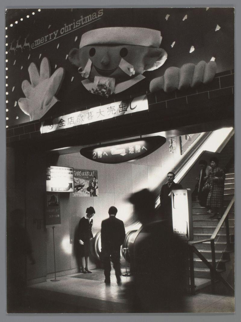 The image is a black and white photograph depicting an interior scene, likely from the 1960s or earlier based on its style. It appears to be taken inside a public building such as a department store or shopping center during Christmas season, indicated by the festive decorations including reindeer silhouettes and snowflakes in the upper left corner with "merry christmas" written above them.
Several people are visible within the space; two individuals can be seen descending on an escalator towards the lower part of the image. Another person is standing at a ticket booth or information desk, while several others appear to be walking through the area. The environment includes signage in Japanese characters, suggesting that this scene takes place in Japan.
The lighting creates a contrast between illuminated areas and darker surroundings, with artificial light sources casting shadows on the subjects and architectural features. Overall, it captures a moment of daily life within an urban se