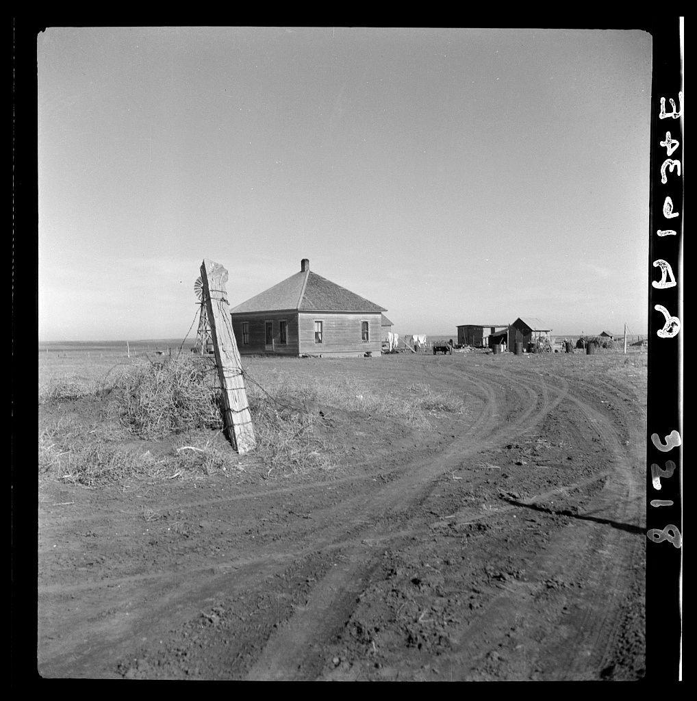 This black and white photograph depicts a rural landscape with an old, weathered wooden house at the center. A large pole stands on one side of the property with what appears to be remnants of windmill blades attached to it. The surrounding area is desolate, featuring open fields that suggest a prairie or plains environment without any trees in sight. In the background, several smaller structures are visible along with scattered household items and livestock, hinting at farming activity. A dirt road winds through the foreground leading away from the house towards these additional buildings. There's clear evidence of human habitation despite its isolated appearance; however, no people can be seen within this frame. The photo is marked with text indicating it belongs to a series or collection by photographer Dorothea Lange and notes that these individuals are expected to be resettled as their land will revert back to cattle range usage.