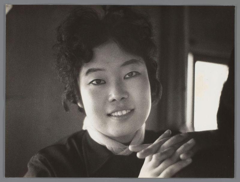 The image is a black and white photograph featuring an individual with shoulder-length curly hair, smiling at the camera. The person appears to be wearing dark clothing with a light-colored collar that suggests layered garments or accessories such as a scarf or neckerchief. In the background, there's a faint reflection of what seems like another figure standing near a window. The overall lighting is soft and diffused, creating gentle shadows on the subject’s face and hands. This photograph evokes an intimate atmosphere with its close-up framing and use of negative space to emphasize the person in focus.