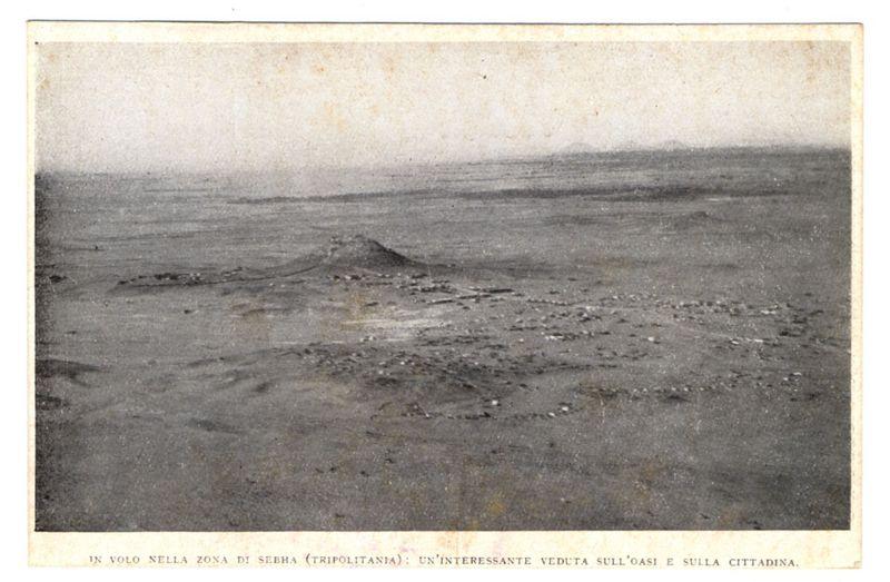 The image depicts a desolate, arid landscape that appears to be taken from an aerial perspective. The terrain is flat with scattered patches of rocky outcroppings and some minor mounds or hills in the distance under a hazy sky suggesting heat haze or atmospheric conditions typical of desert environments.

In black-and-white, it showcases what seems like ancient ruins on sand dunes, possibly indicating historical significance to an oasis region. The text at the bottom indicates that this photo is part of "IN VOLO NELLA ZONA DI SEBHA (TRIPOLITANIA) : UN'INTERESSANTE VEDUTA SULL'OASI E SULLA CITTADINA," which translates from Italian to English as “In flight over the Sebha area in Tripolitania: An interesting view of the oasis and town.”

The text suggests that this image is part of a collection or publication about aerial views, specifically focusing on an area known for its sand dunes (oasis) and towns. The condition of the photograph indicates it might be quite old, likely dating fr