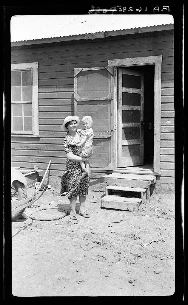 The image is a black and white photograph depicting an outdoor rural scene. A woman, presumably in her late twenties or early thirties based on appearance, stands holding a toddler with both arms around the child's body. She wears a patterned dress, knee-length socks, open-toe sandals, and holds a hat in one hand. The child appears to be under two years old, wearing light-colored clothing suitable for warm weather.

They stand before an older wooden structure that seems worn, possibly indicating age or economic strain on the building's upkeep. To their left is a wheelbarrow filled with various tools and objects indicative of farmwork—rakes, shovels, perhaps even gardening items—and in front lies what looks like a small garden patch.

The ground appears dry and dusty, suggesting an arid climate consistent with rural areas during certain seasons or periods post-drought. There are no other people visible in the immediate vicinity, focusing attention on this duo's moment captured withi