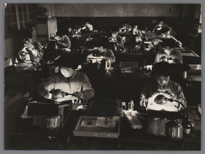 The image is a black and white photograph depicting an industrial setting, likely from the mid-20th century. It shows several individuals working at individual stations equipped with various mechanical devices that suggest they are engaged in precision tasks such as assembly or repair work on small components.

Each worker has their own workstation, which includes what appears to be microscopes or magnifying glasses and other tools necessary for detailed manual labor. The workers are focused intently on the task before them, suggesting a high level of concentration required by their job. In the background, more individuals can be seen performing similar tasks in rows that stretch back into the depths of the room.

The lighting is dim with patches illuminating specific work areas due to overhead artificial light sources or natural daylight coming through unseen windows. The overall atmosphere conveys an environment dedicated to meticulous and repetitive labor typical of a factory se