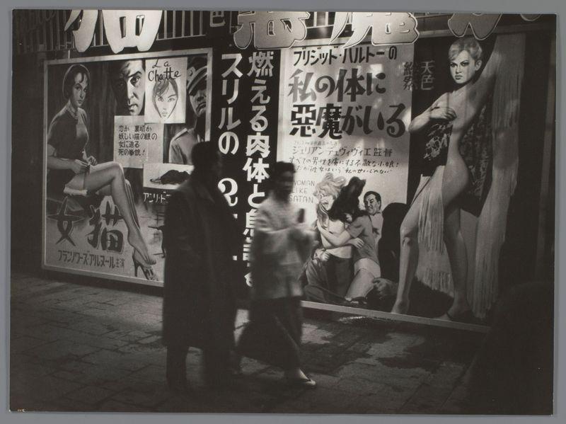 The image is a black and white photograph that captures two people walking on what appears to be a city street. They are in motion, with their bodies slightly blurred due to movement, which gives the impression of a candid moment being captured.

In the background, there's an illuminated billboard featuring large posters of various advertisement images. The text suggests these could be Japanese advertisements for films or products related to entertainment and lifestyle. There is also some English writing on one poster that says "La Chet" with other words beneath it in smaller font, which are not fully legible.

The lighting emphasizes the contrast between the dark street pavement and the bright billboard signs above. The overall atmosphere of the photo suggests a vibrant nightlife or commercial district.