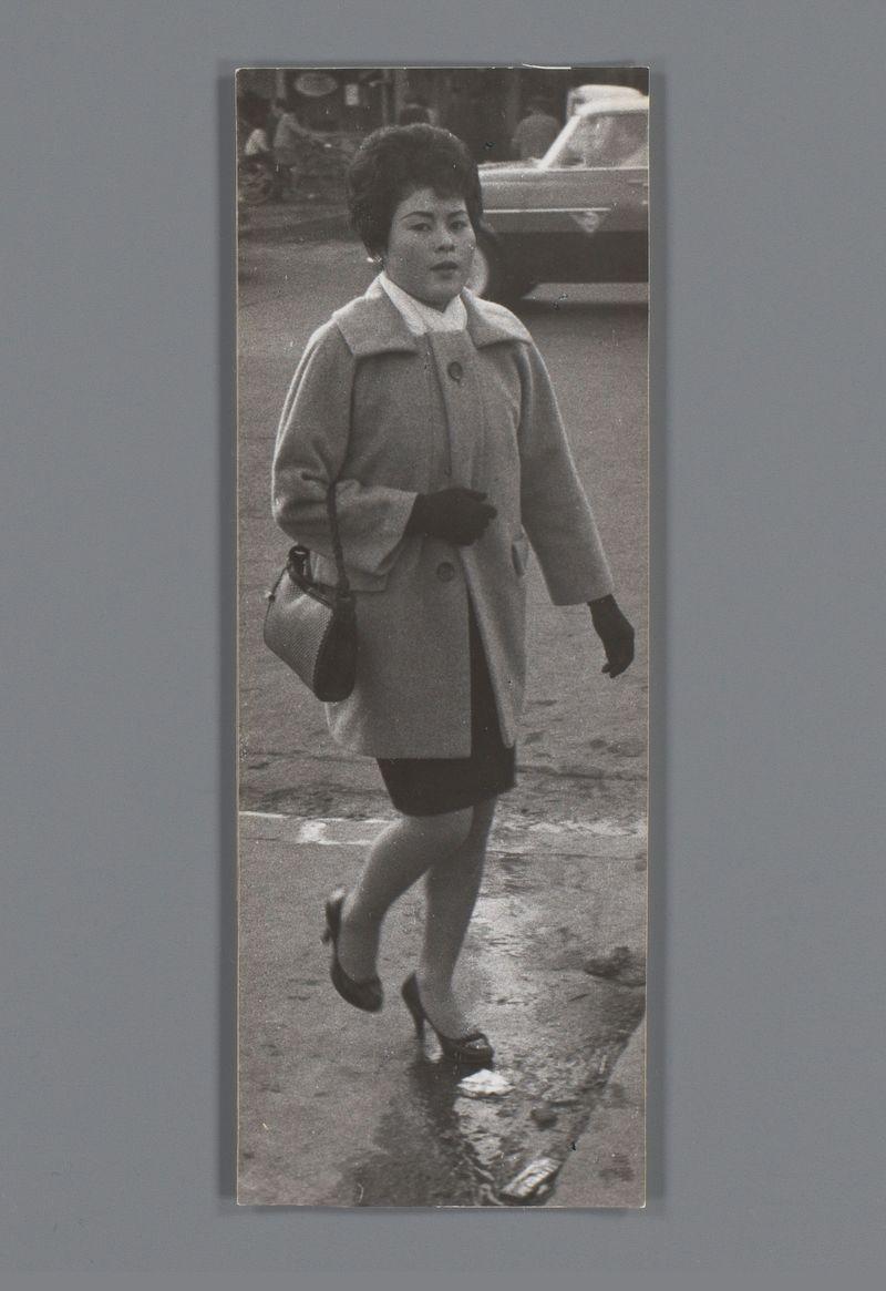The image is a black and white photograph of an East Asian woman walking on what appears to be a wet street. She's wearing a light-colored coat, dark gloves, high-heeled shoes, and carries a small bag over her shoulder. The setting seems urban with reflections indicating the presence of water on the ground, possibly from rain or recent cleaning. In the background, there are blurred images suggesting vehicles and buildings which give an impression of city life. There is no discernible text within the image itself that provides context beyond what has been described visually.

Based on additional information found at Onbekende vrouw op straat in Japan - Ed van der Elsken (1959-60), this photograph may have cultural significance as it captures a moment of everyday life, possibly reflecting societal norms or changes during the mid to late 20th century. The work is attributed to renowned Dutch photographer Édouard "Ed" van der Elsken known for his documentary-style street photography that