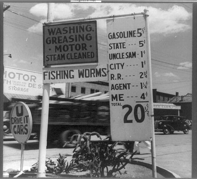 The black and white photograph captures a busy urban scene, likely from the mid-20th century given its monochrome nature. Dominating the foreground is an array of vintage signs advertising various services in bold, capitalized fonts: "WASHING G REASING M OTOR STEAM CLEANED," suggesting steam-powered motor washing; "FISHING WORMS" alongside a phone number for storage and sales indicates a nearby business selling fishing supplies. Another sign announces gasoline prices with rates listed at 5¢, State $1-3¢, Uncle Sam (presumably for government vehicles) $1¢ -2½¢ city R.R.$1¢ to agent 4⅛¢ per gallon; the "ME" cost is humorously priced at "$20." In the background sits a steam engine or locomotive parked on tracks next to an asphalt road, hinting at nearby industrial activity. The presence of cars and palm trees suggests a warm climate, possibly in California, with the caption mentioning Santa Fe, New Mexico as additional context for this image's provenance.