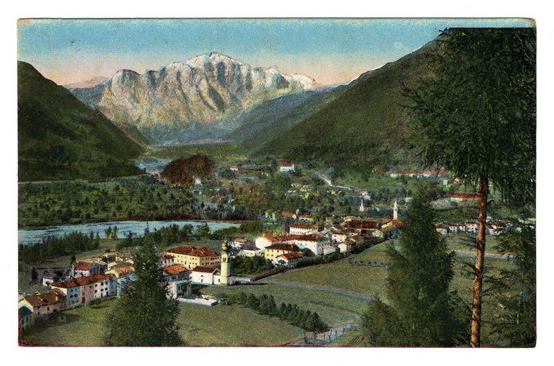 The image depicts a picturesque landscape featuring rolling green hills with dense forestation on the right. In the midground, there's a quaint village nestled among these hills. The architecture suggests European influence, possibly Italian or Alpine in style, characterized by small to medium-sized buildings with red-tiled roofs and pale-colored facades. One prominent structure appears to be a church or large public building due to its size and distinctive design.

A river meanders through the valley below, reflecting sunlight and highlighting various shades of blue and green along its surface. The village is surrounded on one side by what seems like agricultural fields with patches of brown earth visible amidst clusters of crops.

Dominating the background are majestic mountains that rise steeply from behind the hills into a clear sky. These peaks appear to be partially covered in snow, suggesting they could reach considerable altitudes and possibly have seasonal effects such as 