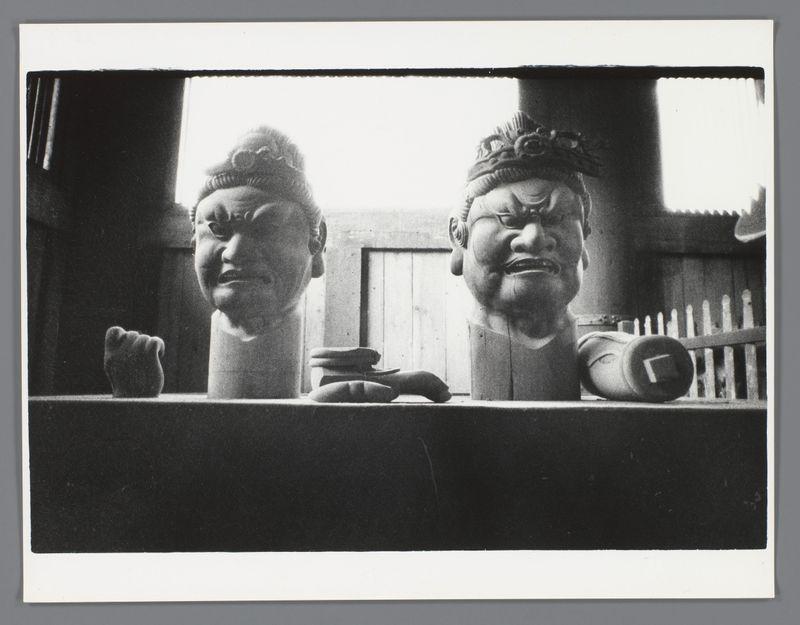 The image depicts a black and white photograph featuring two prominent statues with stern expressions. The statue on the left appears to be wearing traditional headgear, possibly indicative of an ancient figure or deity in Asian cultures, while the one on the right has exaggerated facial features resembling a mask commonly used in theatrical performances. They are set against a backdrop that suggests an interior space, potentially a shrine or museum setting, with wooden elements and natural light filtering through what could be openings above them.

Below these statues is a pedestal upon which various objects rest, including what seems to be traditional footwear and possibly ceremonial items like bracelets or rings of gold. The composition evokes themes of cultural heritage, ritualistic practices, or historical commemoration, captured in monochrome tones that enhance the gravity and solemnity of the scene.